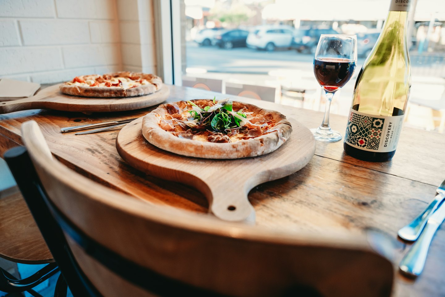 Two pizzas on wooden peels with wine on a table.