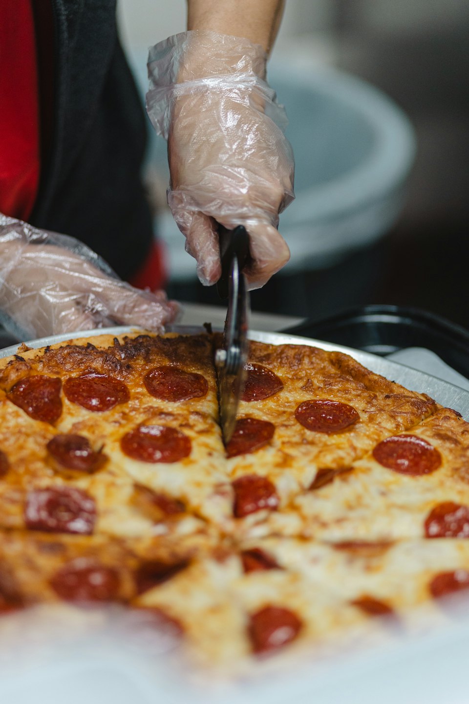 Hands wearing plastic gloves slice a pepperoni pizza with a cutter.